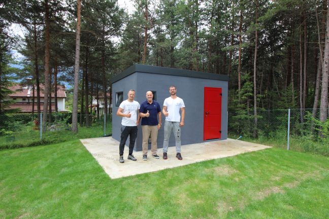 Spokojný zákazník pred záhradným domčekom v tmavo-sivej omietke s červenými dverami Hormann