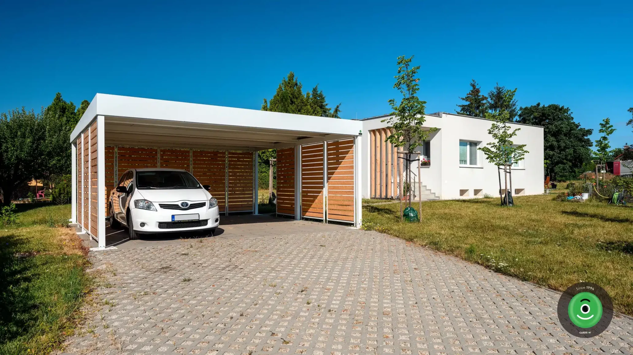 Biely prístrešok GARDEON s bielym autom vo vnútri na záhrade.