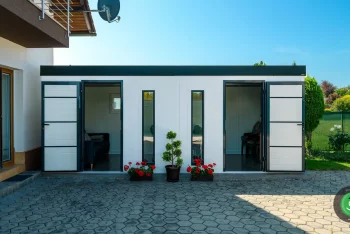 Biely záhradný domček s pultovou strechou s čiernou atikou pri dome.