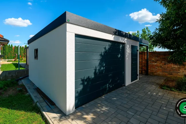 Biela montovaná garáž s tmavosivou bránou a dverami, elegantný vzhľad na dvore rodinného domu.